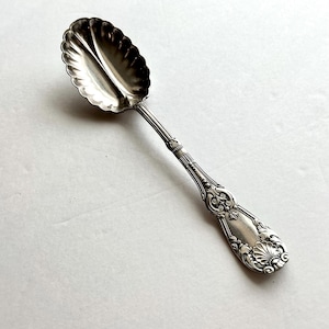 May include: A silver serving spoon with a scalloped bowl and ornate handle. The spoon's bowl has a fluted design, and the handle features detailed floral and scrollwork patterns. The spoon is likely made of sterling silver.