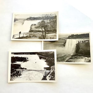 May include: Three vintage black and white postcards featuring Niagara Falls. One postcard shows the U.S. view from Luna Island, another shows Goat Island, and the third shows the Niagara River and a bridge. The postcards are arranged on a white surface.