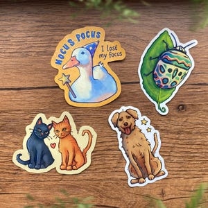 Op de afbeelding: Vier gestanste stickers met een witte eend in een tovenaarshoed met de tekst HOCUS POCUS I lost my focus, een kleurrijke kever op een groen blad, een zwarte en een oranje kat met een klein hartje, en een beige hond met twee sterretjes.