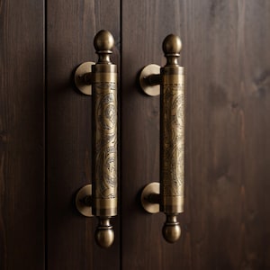 May include: Two vertical door handles made of antique brass with a cylindrical shape and spherical finials at the top and bottom. The central section of each handle features an intricate, engraved pattern of swirling lines and geometric shapes.