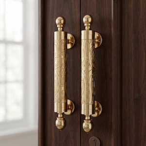 May include: Two vertical brass door handles with a polished gold finish and intricate engraved patterns. Each handle features a cylindrical center with rounded finials at the top and bottom, mounted to a dark wood door with circular metal plates.