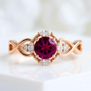 May include: A rose gold ring featuring a large, round, deep pink center stone. The center stone is surrounded by four marquise-cut white stones and two smaller round white stones. The band has an intricate, open, twisted metal design.