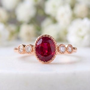 Peut inclure: Une bague en or rose ornée d'un rubis rouge profond taille ovale serti dans une bordure millegrain. L'anneau est agrémenté de chaque côté par deux petits diamants blancs ronds taille brillant, chacun serti dans un cadre millegrain assorti.