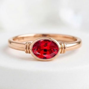 May include: A rose gold ring featuring a central oval-cut vibrant red gemstone. The band is smooth and polished, with two small vertical ridges on each side of the setting that secure the stone in place, creating a clean and balanced appearance.