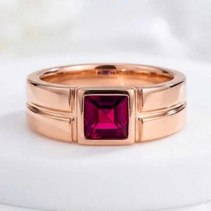 Könnte beinhalten: Ein Ring aus Roségold mit einem quadratisch geschliffenen, tiefroten Edelstein in der Mitte. Die Ringschiene hat eine glatte, polierte Oberfläche mit einem breiten, flachen Profil und einem markanten, gestuften Design um die quadratische Fassung.