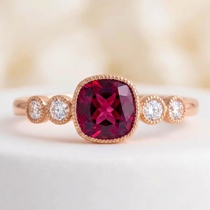 May include: A rose gold ring featuring a central cushion-cut deep red gemstone set in a milgrain bezel. The band is accented on each side by two round brilliant-cut white diamonds, each held in a circular milgrain setting, creating a symmetrical design.