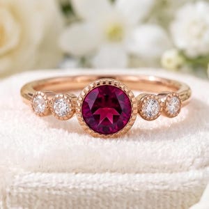 May include: A rose gold ring featuring a central round-cut deep red gemstone in a milgrain bezel setting, flanked by two smaller round white diamonds on each side, also set in milgrain-detailed rose gold bezels on a polished rose gold band.