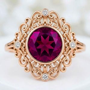May include: A rose gold ring featuring a large, round, deep magenta center stone surrounded by an intricate filigree metal frame. Four small, clear, round-cut accent stones are set at the top, bottom, left, and right of the central setting.