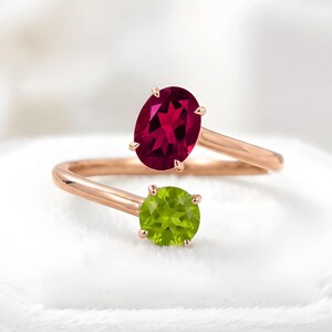 May include: A bypass ring crafted from rose-colored metal featuring two prong-set gemstones. One stone is a deep red oval-cut garnet, and the other is a vibrant lime green round-cut peridot, both positioned at the open ends of the polished band.