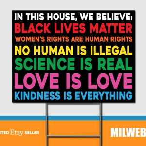 May include: A black yard sign with the text "IN THIS HOUSE, WE BELIEVE: BLACK LIVES MATTER, WOMEN'S RIGHTS ARE HUMAN RIGHTS, NO HUMAN IS ILLEGAL, SCIENCE IS REAL, LOVE IS LOVE, KINDNESS IS EVERYTHING." The sign is supported by a metal frame.