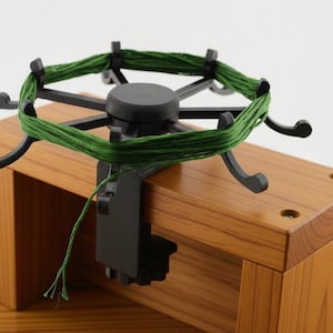 May include: A black plastic yarn swift with a circular frame and eight curved arms, mounted onto a light brown wooden block. Green yarn is wound around the perimeter of the arms, and a single strand of green yarn hangs down from the device.