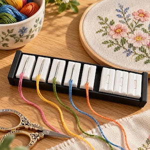 May include: A black rectangular needle organizer with eight white magnetic slots, five of which hold silver needles threaded with pink, yellow, green, blue, and orange embroidery floss. The organizer sits near a pair of ornate metal scissors.