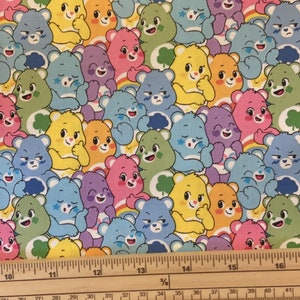 May include: A colorful fabric with a repeating pattern of Care Bears. The bears are all smiling and have different colored fur, including blue, yellow, pink, green, and purple. The fabric is measured with a ruler in inches.