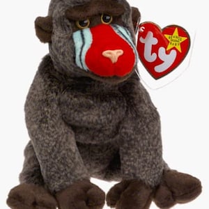May include: A brown and gray stuffed monkey with a red face and blue markings around its eyes. The monkey is sitting with its legs crossed and has a Ty Beanie Baby tag attached to its ear.
