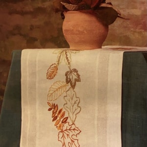 May include: A white linen table runner with an embroidered design of autumn leaves in shades of orange, brown, and green. The runner is on a dark green surface and a terracotta pot with a red flower is in the background.