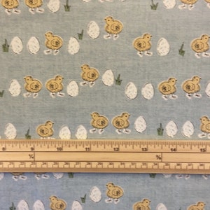 May include: A blue fabric with a repeating pattern of yellow chicks and white eggs. A wooden ruler is placed on top of the fabric.