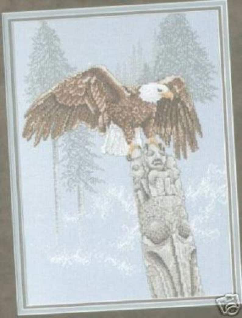 May include: A brown and white bald eagle with its wings spread wide, perched on a weathered totem pole. The eagle is facing to the right, and the totem pole is carved with a face. The background is a snowy forest scene.