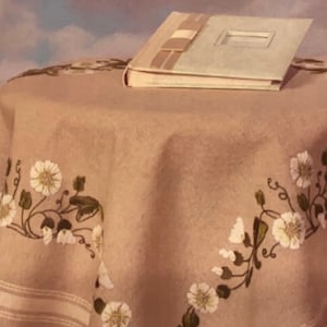 May include: A beige tablecloth with embroidered white flowers and green leaves. A white photo album with a beige ribbon is on top of the tablecloth.