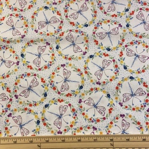 May include: White fabric with a repeating pattern of blue dragonflies surrounded by colourful floral wreaths. The fabric is measured with a wooden ruler.