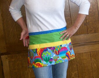 Peut inclure: Un tablier coloré avec un motif de montgolfières. Le tablier comporte une bande bleue, verte et jaune en haut, et un motif vibrant de montgolfières de différentes couleurs. La personne porte un t-shirt blanc à manches longues et un jean bleu.