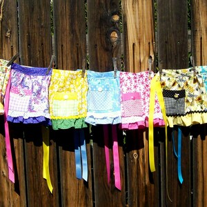 May include: A collection of colorful, handmade aprons hanging on a clothesline against a wooden fence. Each apron features a unique pattern, including floral, polka dots, and patchwork designs, with contrasting trim and ribbons. The aprons are in various colors, including pink, yellow, blue, and purple.