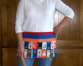 Peut inclure: Un demi-tablier coloré avec un motif de casiers scolaires. Le tablier présente une ceinture rouge, une section à pois bleus et une section inférieure avec un motif répété de casiers de différentes couleurs. Le tablier est porté sur un haut blanc à manches longues et un jean bleu.