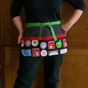 Peut inclure: Un tablier noir avec une ceinture et des liens verts, orn&eacute; de pois et d'une bordure rouge. Le tablier est orn&eacute; de divers &eacute;cussons color&eacute;s et de texte, dont les mots "Teacher" et "Great Teacher".