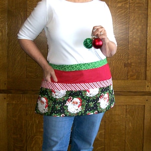 May include: A half apron featuring horizontal bands of green fabric, red fabric with a white dot pattern, and red and white diagonal stripes, finished with a bottom panel displaying a black background with Santa Claus and holly.