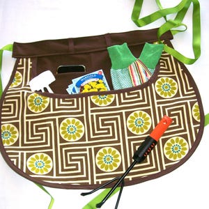 May include: Brown gardening apron with a geometric floral pattern, green ribbon ties, and multiple pockets. The pockets contain gardening gloves, a small hand rake, a white tool, and seed packets. The apron is designed for gardening.