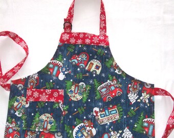 Puede incluir: Delantal navideño con fondo azul oscuro y diseños de coches rojos, muñecos de nieve y caravanas. Cuenta con un bolsillo y ribetes rojos con motivos de copos de nieve blancos. El delantal tiene una correa para el cuello y lazos en la cintura rojos.