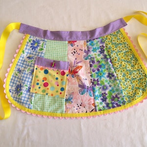 Puede incluir: Un delantal de patchwork con una variedad de telas florales y estampadas. El delantal presenta una cinta amarilla, un bolsillo con lunares coloridos y un borde festoneado con ribete rosa. El delantal está diseñado para uso en la cocina.