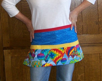 Peut inclure: Un tablier coloré avec un motif de montgolfières. Le tablier a une bordure rouge à pois, une section centrale bleue à motifs et une bande jaune. La partie inférieure présente des montgolfières vibrantes de différentes couleurs.
