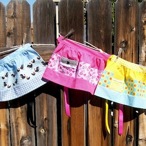 May include: A row of colorful half aprons hanging on a wooden fence. The aprons feature various patterns, including floral, polka dots, and chicken motifs. Each apron has a pocket and ties, with a mix of pink, blue, and yellow hues.
