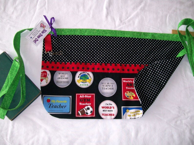 Peut inclure: Tablier de professeur noir avec une bordure et des bretelles vertes. Le tablier pr&eacute;sente un motif &agrave; pois noirs et blancs, un ruban &agrave; pois rouges et des &eacute;cussons avec les mentions "Meilleur professeur du monde" et "Mon professeur pr&eacute;f&eacute;r&eacute;".