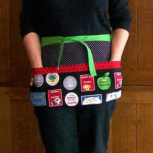 Peut inclure: Un tablier noir avec une section sup&eacute;rieure &agrave; pois verts et blancs et une section inf&eacute;rieure noire. La section inf&eacute;rieure comporte divers &eacute;cussons color&eacute;s avec du texte tel que "Great Teacher" et "World's Best Teacher". Une cravate verte est nou&eacute;e &agrave; la taille.
