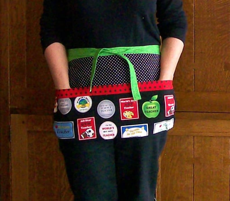 Peut inclure: Un tablier noir avec une section sup&eacute;rieure &agrave; pois verts et blancs et une section inf&eacute;rieure noire. La section inf&eacute;rieure comporte divers &eacute;cussons color&eacute;s avec du texte tel que "Great Teacher" et "World's Best Teacher". Une cravate verte est nou&eacute;e &agrave; la taille.