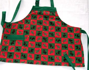 Puede incluir: Un delantal con temática navideña con un patrón de cuadros rojos y verdes. El delantal presenta un diseño repetido de perros Scottie negros y árboles de Navidad. Tiene un bolsillo verde y correas verdes.