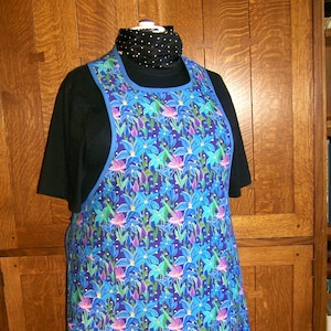 May include: A blue apron with a floral pattern of blue, purple, and green flowers. The apron has a blue trim and is displayed on a mannequin. The background is a wooden cabinet.