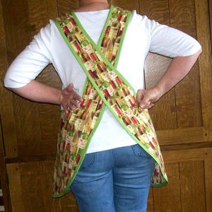 May include: An apron featuring a wine bottle pattern with green trim. The apron is worn over a white shirt and blue jeans. The apron's straps cross in the back. The wine bottle design includes red, white, and green bottles.