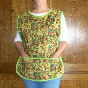 May include: A green apron with a floral pattern featuring red and blue accents. The apron has a green trim and two front pockets. The person is wearing a white long-sleeve shirt and blue jeans.