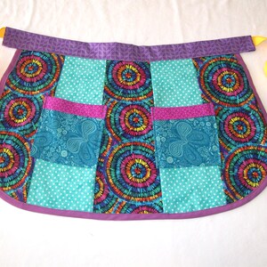 Puede incluir: Un delantal de media cintura colorido con un diseño de patchwork. El delantal presenta una cinturilla y un ribete morados, con lazos amarillos. El cuerpo principal del delantal está formado por cuadrados de tela con estampados turquesa, rosa y arcoíris.