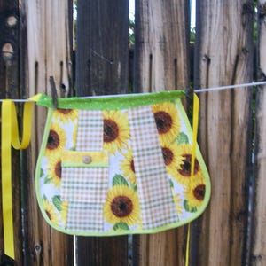 Puede incluir: Delantal hecho a mano con un diseño de girasoles sobre un fondo a cuadros. El delantal tiene un bolsillo con un botón y está ribeteado con tela de lunares verdes. Cintas amarillas cuelgan de los lados. El delantal está colgado en un tendedero.
