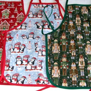 May include: Three Christmas-themed aprons. One red apron features gingerbread houses and figures. Another is light blue with penguins wearing Santa hats. The third is green with nutcracker soldiers.