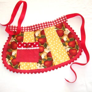 Puede incluir: Delantal hecho a mano con un estampado de manzanas vibrantes sobre un fondo amarillo con lunares blancos. El delantal tiene un borde festoneado rojo, un bolsillo rojo con ribete de encaje blanco y un ribete de cuadros rojos en la parte superior. Lazos de cinta roja completan el diseño.