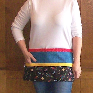 May include: A half apron with a red polka dot band, blue denim section, and a yellow band. The bottom section features a black fabric with a colorful space-themed print of rockets, planets, and stars. The apron is worn over a white long-sleeved shirt and blue jeans.