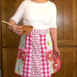 May include: A patchwork apron with a pink and white gingham pattern, floral pockets, and a pink trim. The apron is worn over a white long-sleeved shirt. A wooden bowl and spoon are held in the person's hand.