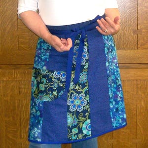 Könnte beinhalten: Eine blaue Schürze mit floralem Muster und Wickeldesign. Die Schürze weist eine Vielzahl von Blautönen auf, darunter ein einfarbiges blaues Panel und Abschnitte mit Blumen- und Paisley-Drucken. Sie hat einen Bindverschluss an der Taille.