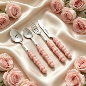 May include: A set of four stainless steel utensils including two spoons, a fork, and a knife with light pink, spherical, beaded handles. The utensils are engraved with the letters C, A, L&A, and the text Lucas&Audrey, arranged on a satin fabric.