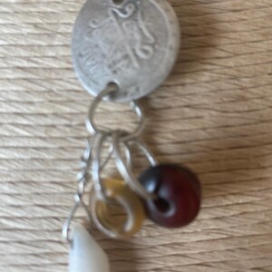 May include: A silver charm necklace featuring a vintage coin and several dangling charms. The charms include a white teardrop, a yellow disc, and a dark red bead. The necklace is displayed against a textured, light brown background.