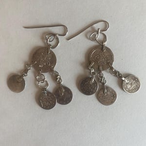 May include: Silver-toned dangle earrings featuring coin-shaped charms. Each earring has a larger coin at the top with smaller coins dangling below, connected by small chain links. The earrings have a simple hook closure.